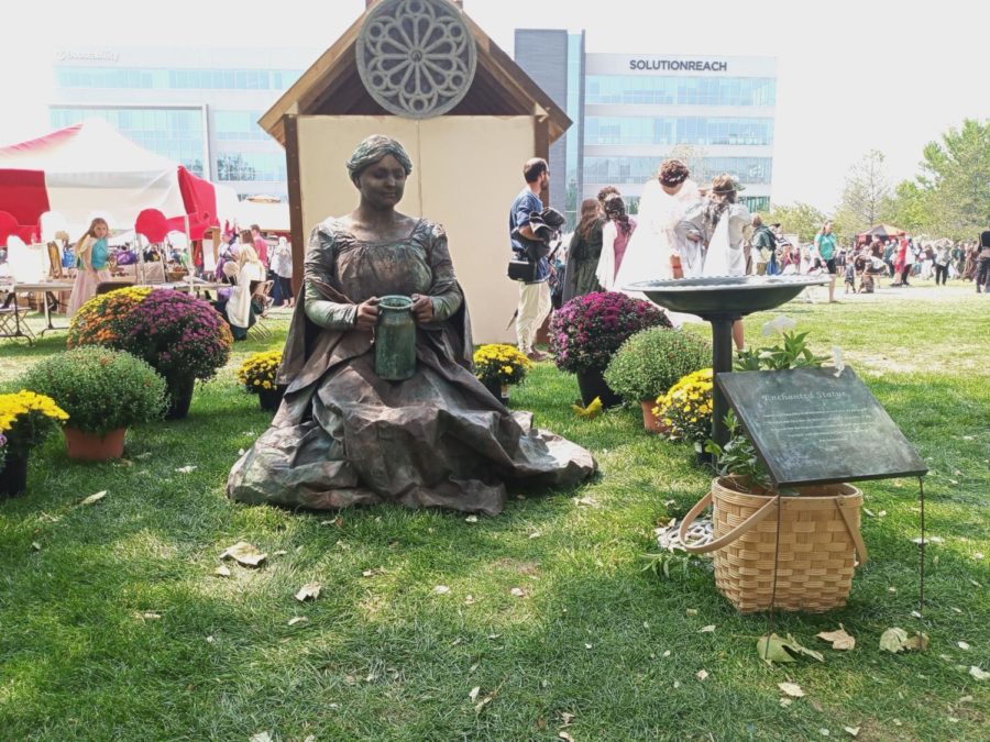 A picture of a living statue. The statue is meant to look like a weaping woman made of aged metal. She sits in grass with a metal pot and a small basket to collect tips