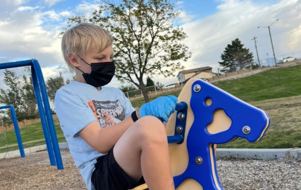 A sad young boy is playing alone at a park with a mask and gloves on to keep himself safe from COVID-19.